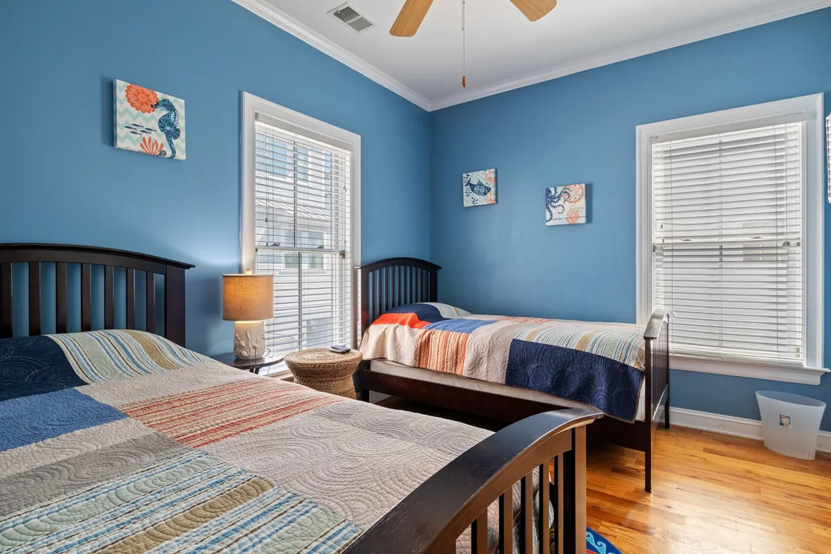 A guest bedroom with two beds dressed in colorful bedspreads, blue walls, and a bedside lamp. The space is cozy and suitable for families or groups, with a mood that encourages stories, laughter, and restful sleep.