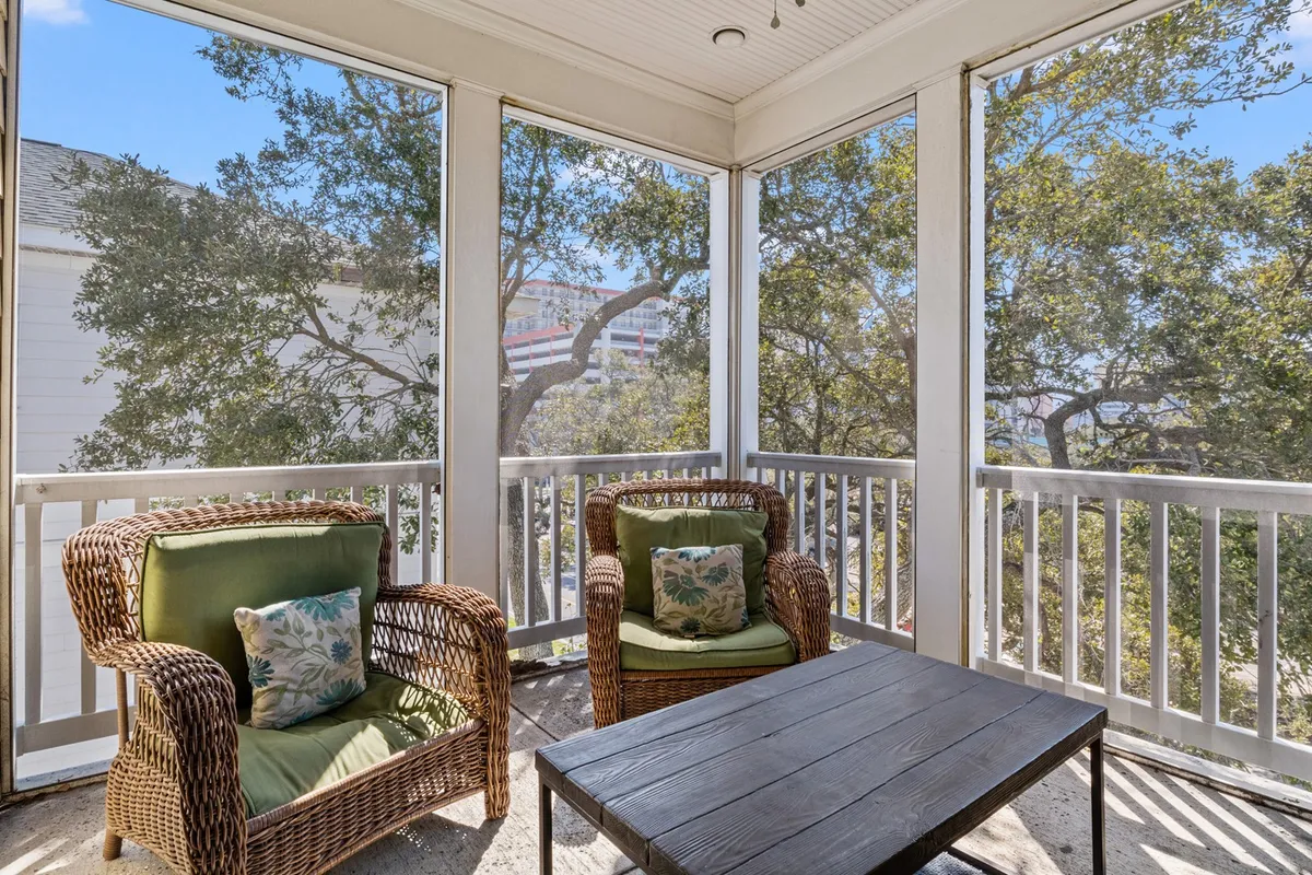 A sunlit sitting area with two wicker chairs and a wooden coffee table situated before large windows, inviting relaxation. The view outside blends nature with indoor tranquility, while the room exudes a cozy, calm atmosphere perfect for unwinding or socializing.