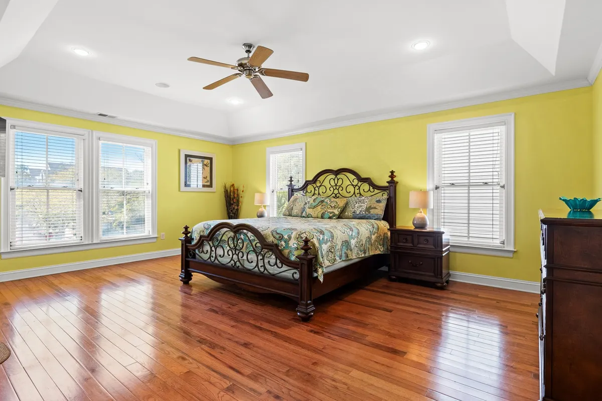 A spacious bedroom featuring an ornate bed with colorful, plush bedding, complemented by wooden furniture and large windows that welcome abundant natural light. The overall ambiance is comfortable and inviting, ideal for unwinding after a day at the beach.
