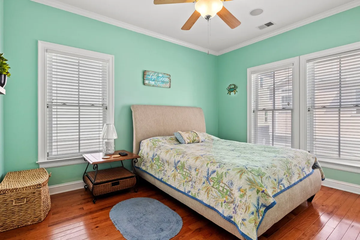 A serene bedroom setting with a bed adorned in floral bedding, mint green walls, windows with blinds, and decorative wall art. The atmosphere is calm, perfect for relaxation, reading, or napping in a peaceful environment.