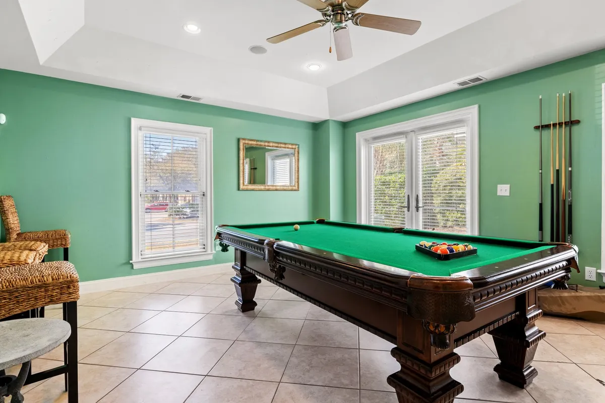 This image captures a lively game room featuring a pool table at the center, surrounded by green walls and ample windows letting in natural light. A cozy seating area is visible where people can relax or socialize, contributing to a fun and casual atmosphere. The scene exudes the spirit of friendly competition and memorable gatherings.