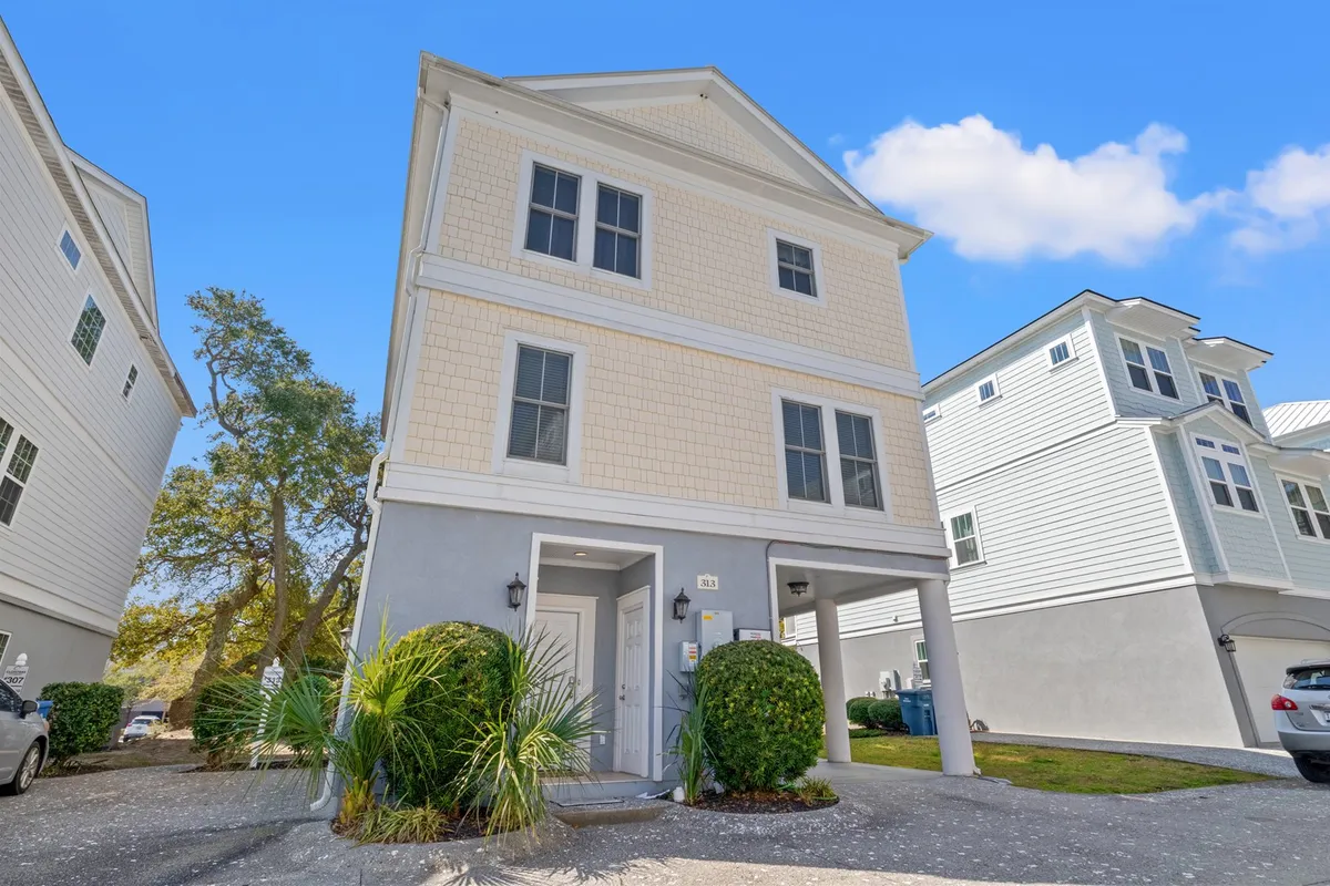 A welcoming three-story house with a landscaped front yard dotted with palm trees and a spacious driveway, hinting at plenty of space for family gatherings and easy parking. The home’s coastal architecture is set against a backdrop of blue skies, capturing the vacation vibe and offering a clear sense of arrival steps from the beach.