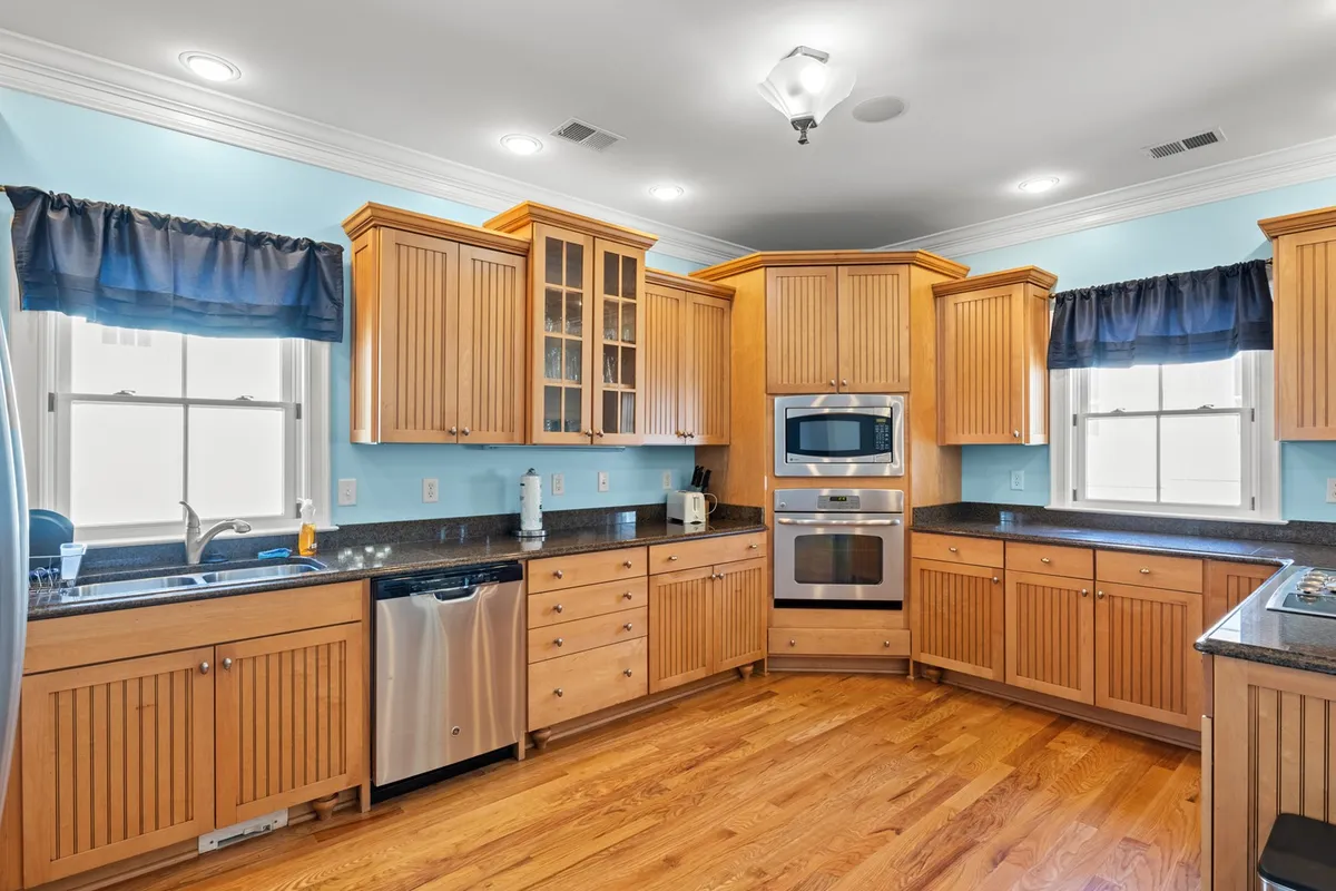 A modern kitchen with wooden cabinetry, sleek black granite countertops, stainless steel appliances, and blue-curtained windows. The space feels bright and welcoming, ready for cooking or entertaining.